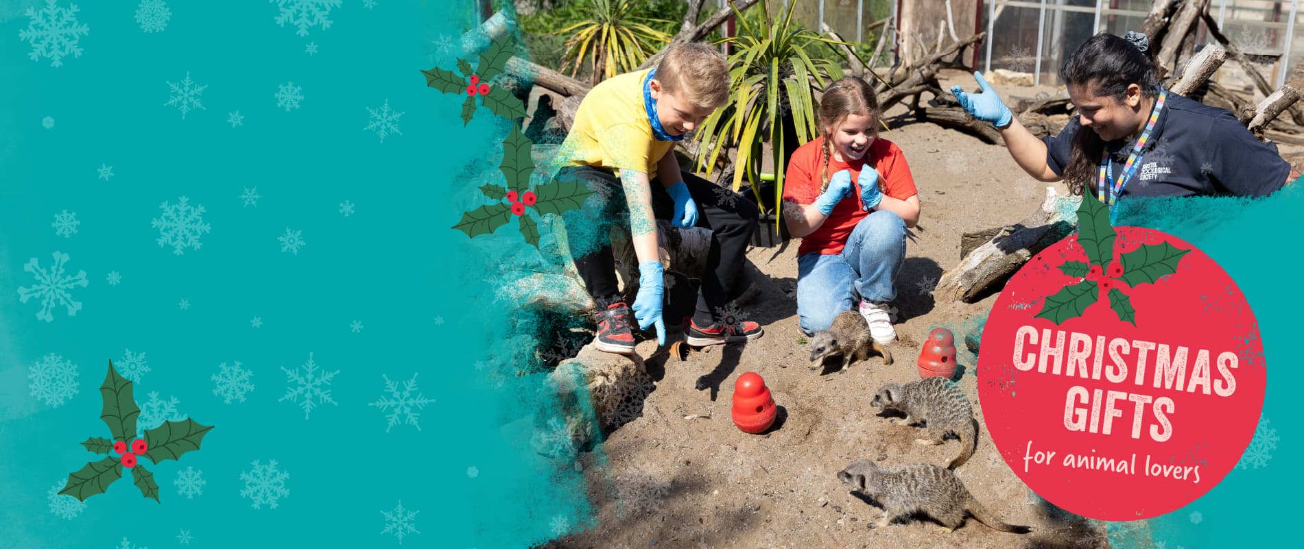 Two children and a zoo staff member crouch in the meerkat habitat. A Christmas Gifts message is overlaid