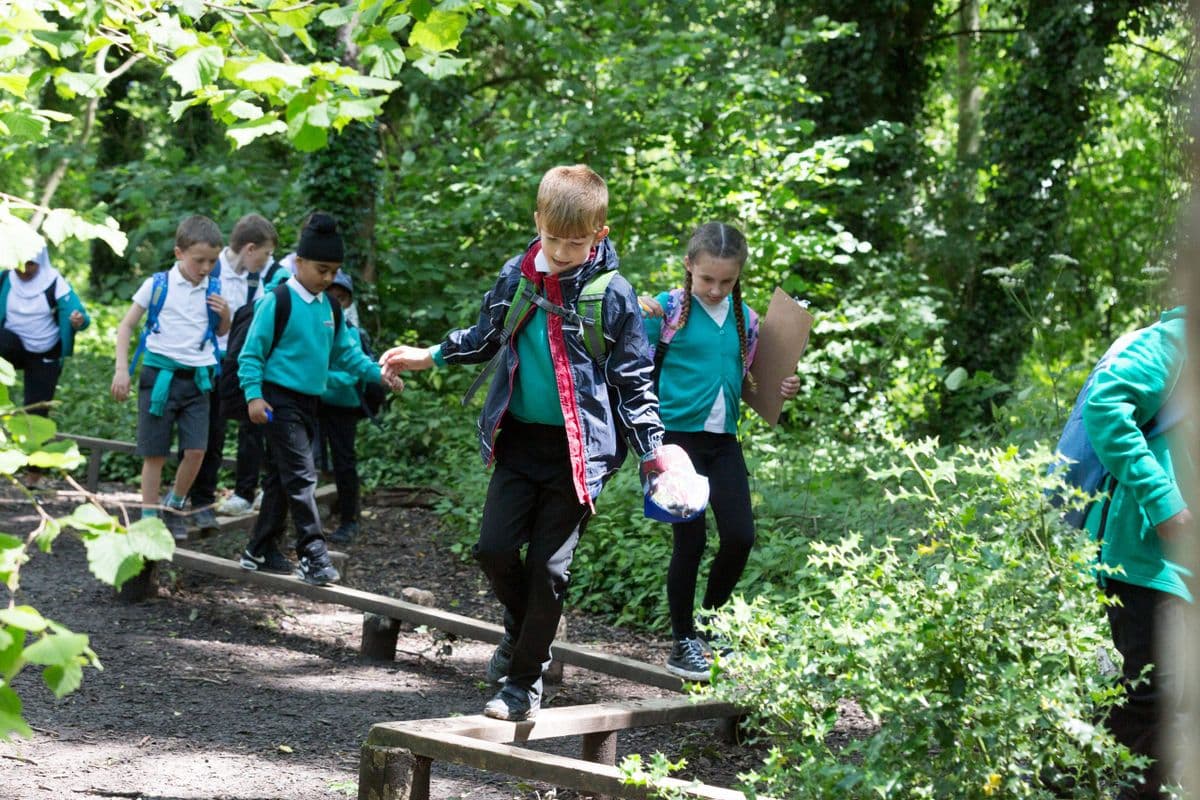 Children in school uniform holding clipboards walk across a small wooden walkway.